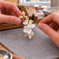 A close-up of hands holding a delicate floral hair pin with white flower petals, gold leaves, and pearl-like centers over a gray work surface in a jewelry-making setting.