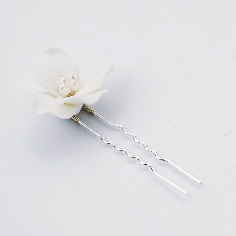 A white flower hair pin with two silver spiral pins and small pearl-like accents in the center, photographed on a clean light gray background.